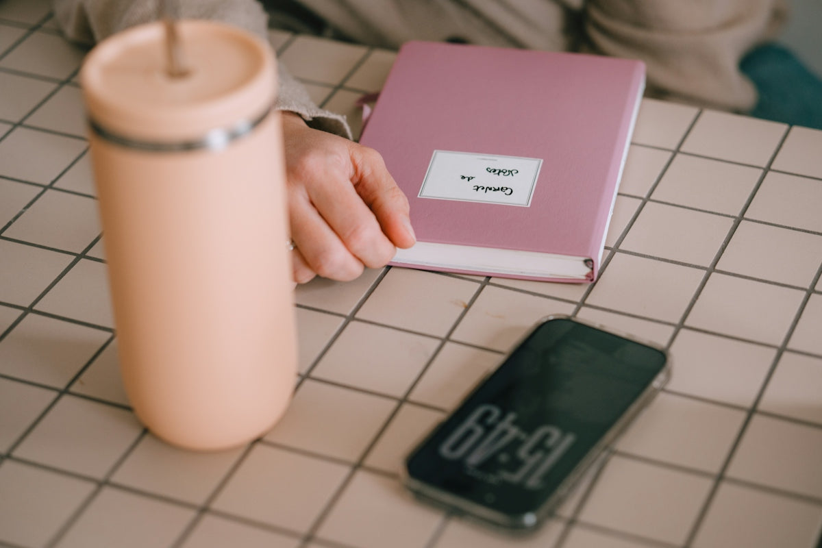 A pink tumbler, book, and phone on a checkered surface